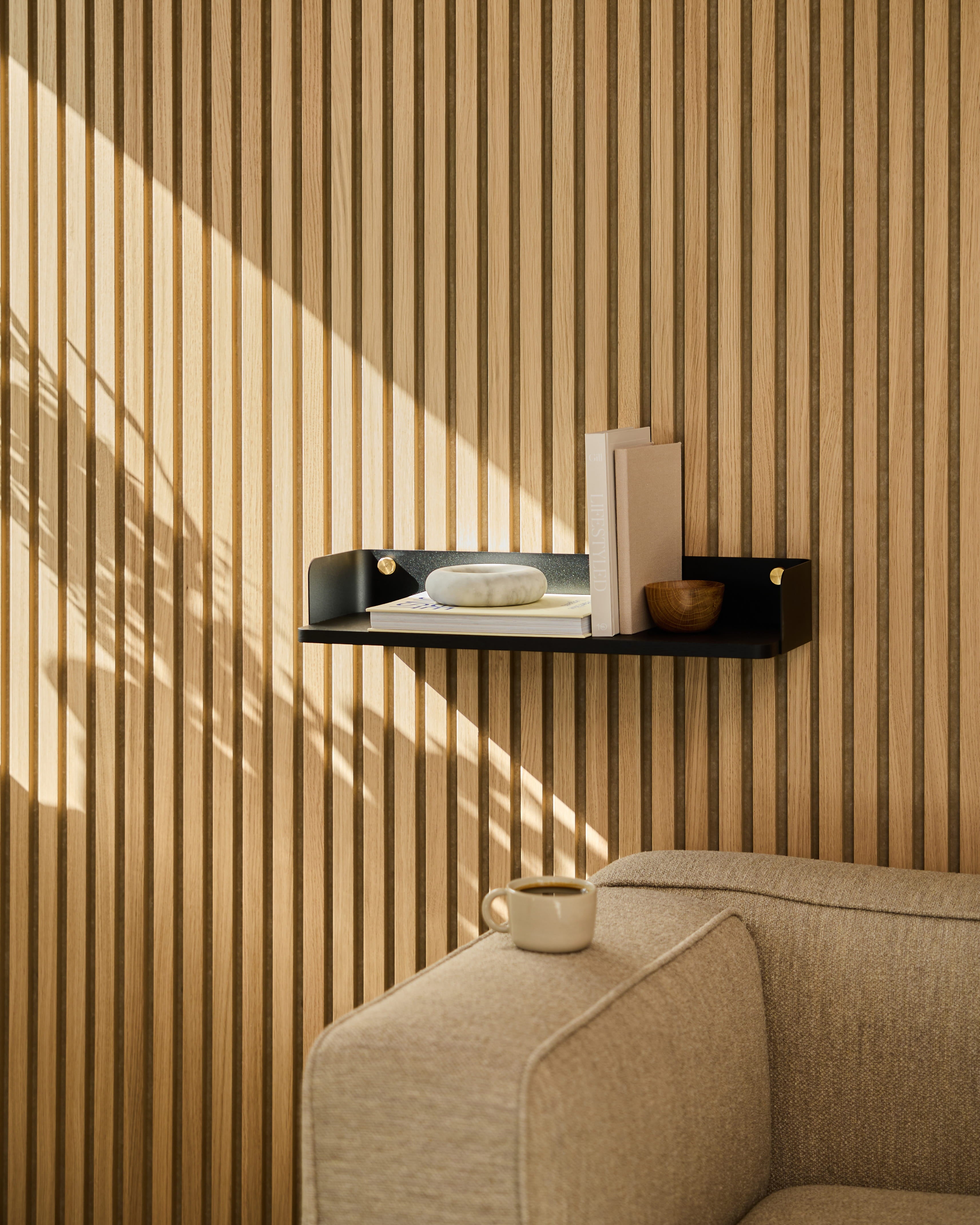 Regał Linn | Bookshelf / Black Oak marki WoodUpp stanowi element dekoracyjny nad beżową sofą. Promienie słoneczne rzucają cienie na ścianę pokrytą drewnianymi panelami akustycznymi w paski.